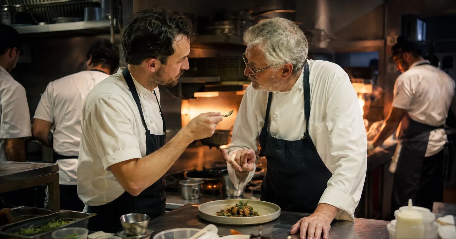 Senior chef offering feedback to younger chef tasting a dish in busy restaurant kitchen